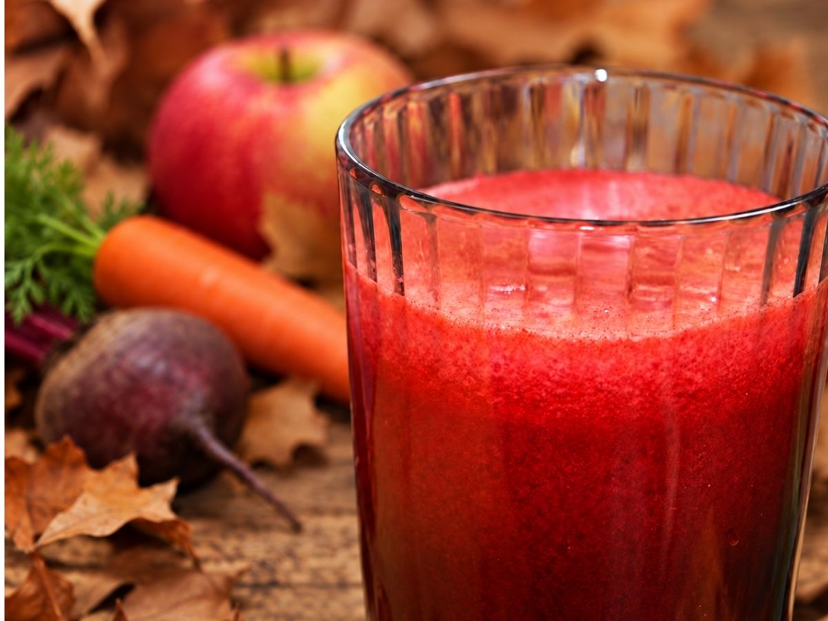 Close-up of apple, beet and carrot juice in a ribbed glass, with a whole beetroot, carrot and apple behind it