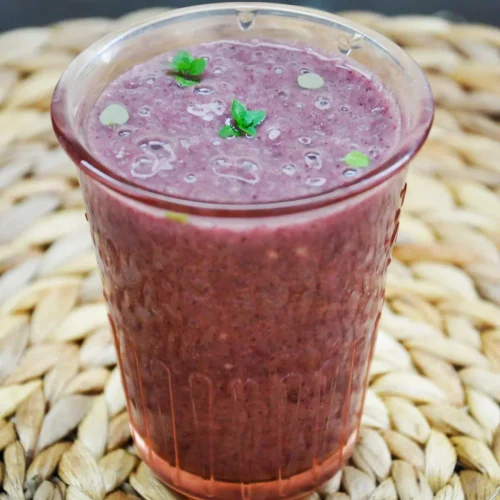 Acai banana smoothie in a tall pink-tinted glass, topped with tiny mint leaves and small bubbles, standing on a round woven placemat against a dark background.