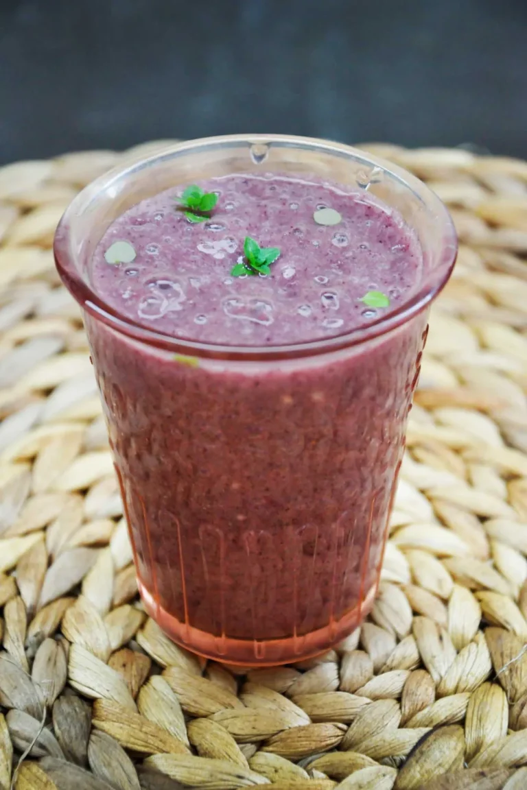 Acai banana smoothie in a tall pink-tinted glass, topped with tiny mint leaves and small bubbles, standing on a round woven placemat against a dark background.
