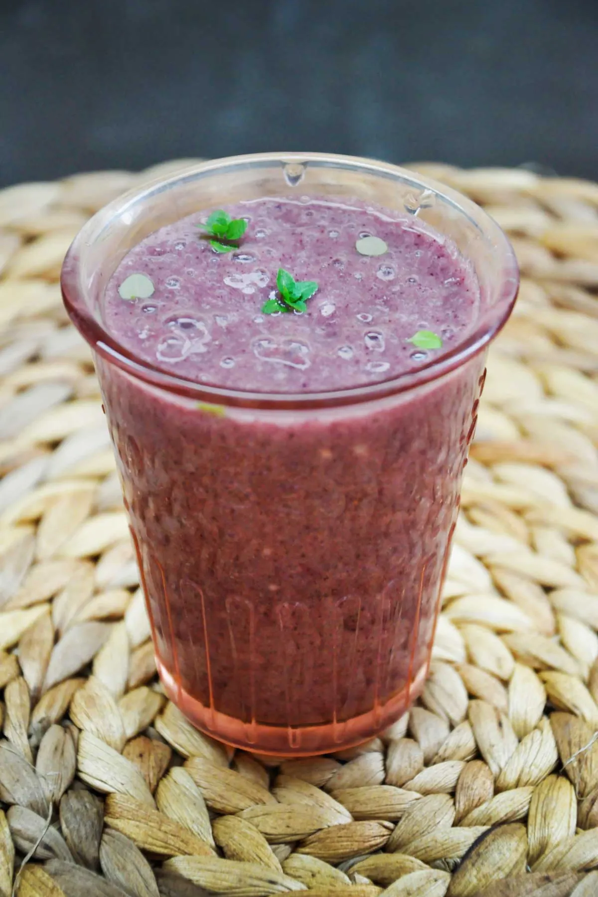 Acai banana smoothie in a tall pink-tinted glass, topped with tiny mint leaves and small bubbles, standing on a round woven placemat against a dark background.