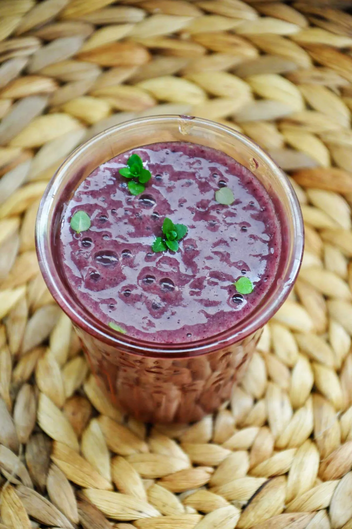 Overhead view of a purple acai berry smoothie in a glass, with mint leaves floating on the bubbly surface, set on a round woven placemat.