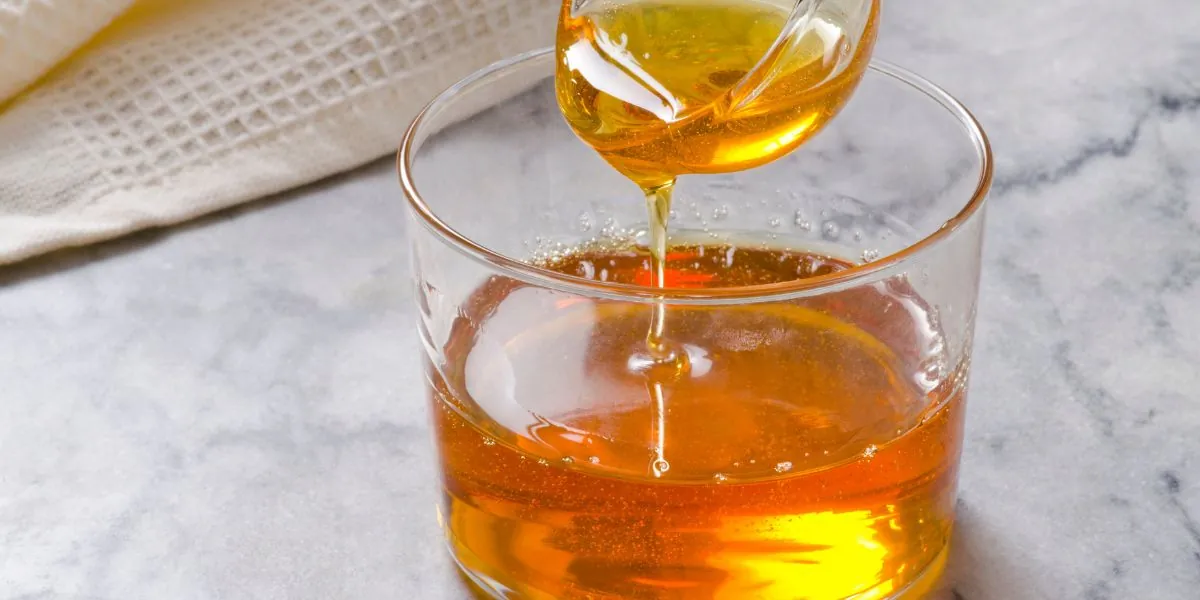 Sirop de agave auriu turnat dintr-o cănuță de sticlă într-un pahar transparent, pe un blat de marmură albă, cu un ștergar de in în spate.