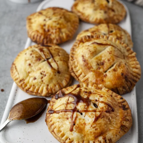 Apple hand pies with a caramel drizzle on a white platter, next to a spoon of caramel sauce