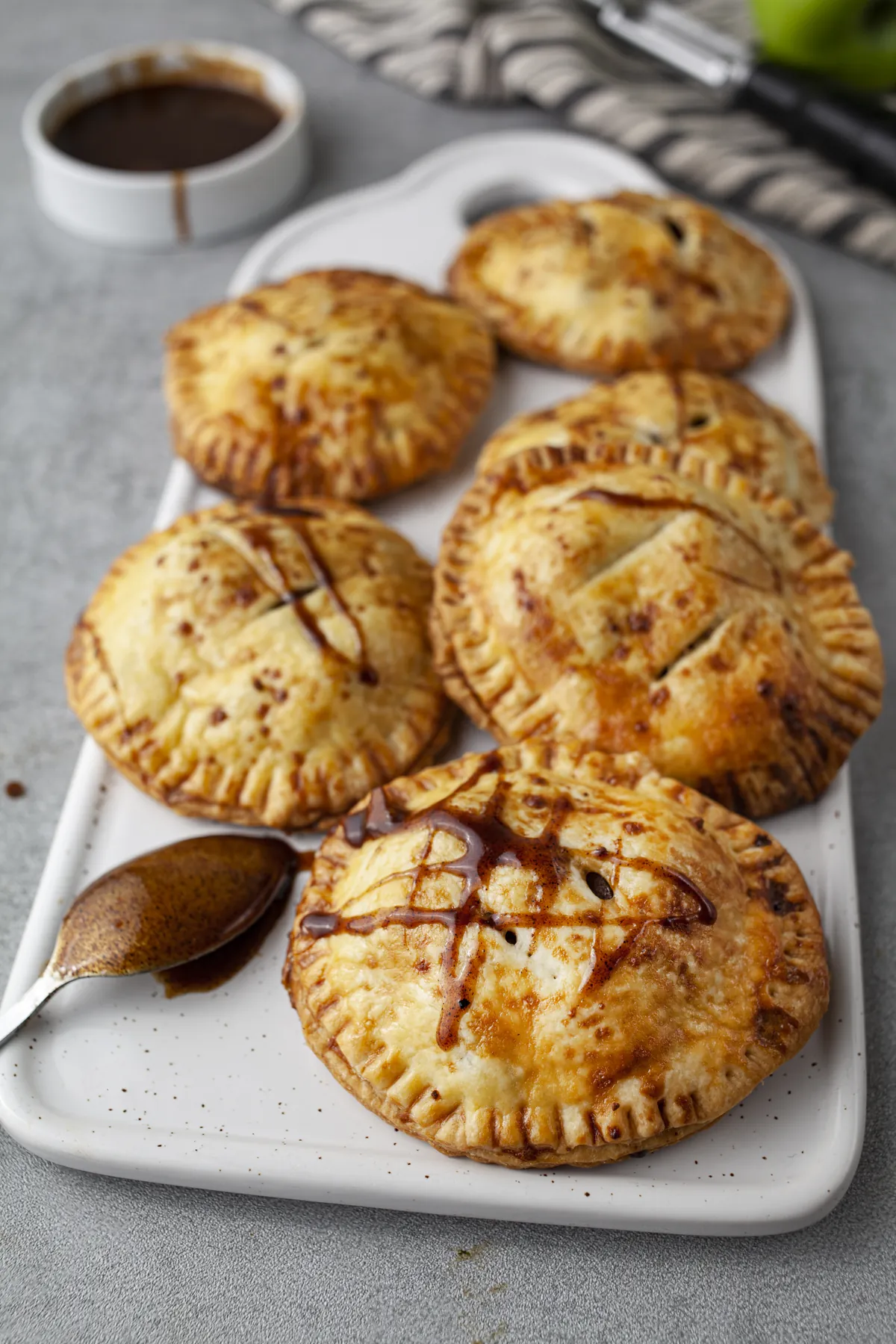 Apple hand pies with a caramel drizzle on a white platter, next to a spoon of caramel sauce