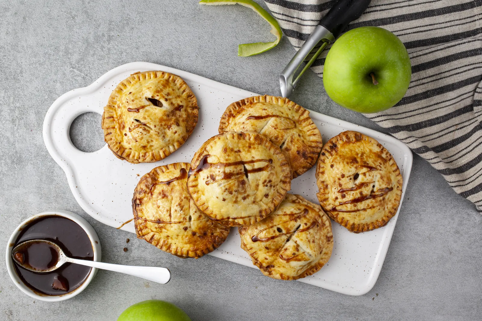 Overhead view of seven baked apple hand pies on a white board, with a small bowl of caramel sauce and a green apple