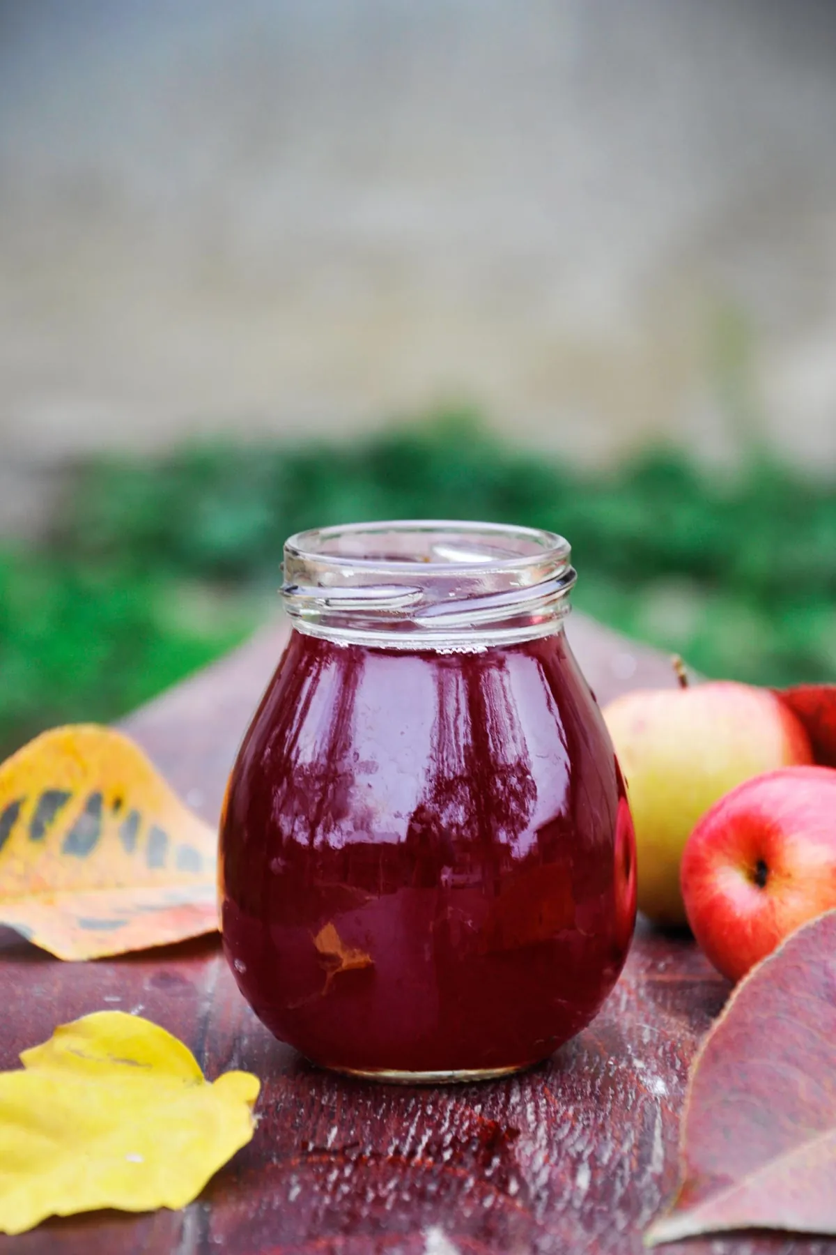 Apple and sour cherry juice in a round glass jar on a weathered red wooden table, with apples and yellow autumn leaves around it and a green garden behind.