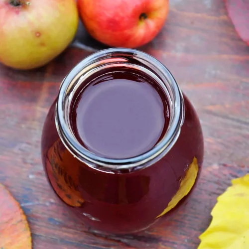 Overhead view of deep ruby apple and sour cherry juice in an open glass jar, with two red and yellow apples and fallen autumn leaves on a wooden table.
