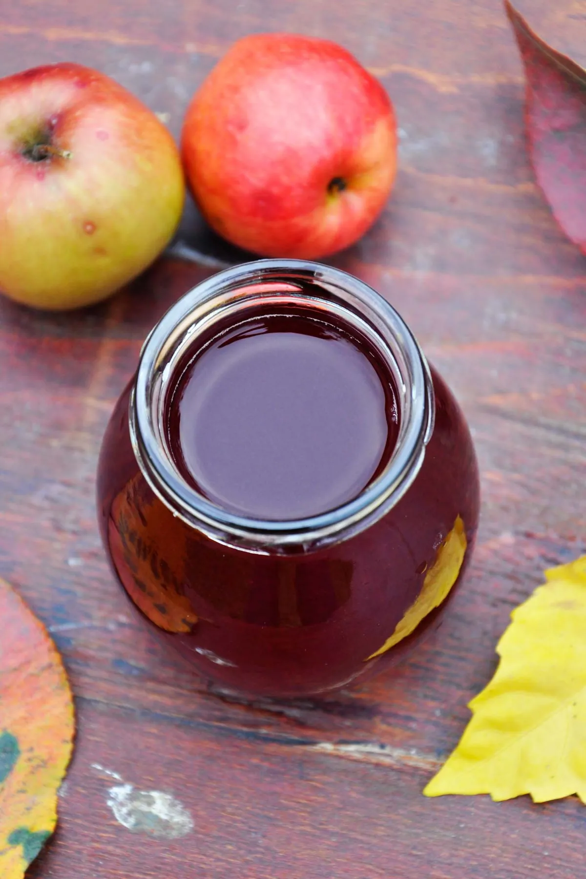 Overhead view of deep ruby apple and sour cherry juice in an open glass jar, with two red and yellow apples and fallen autumn leaves on a wooden table.