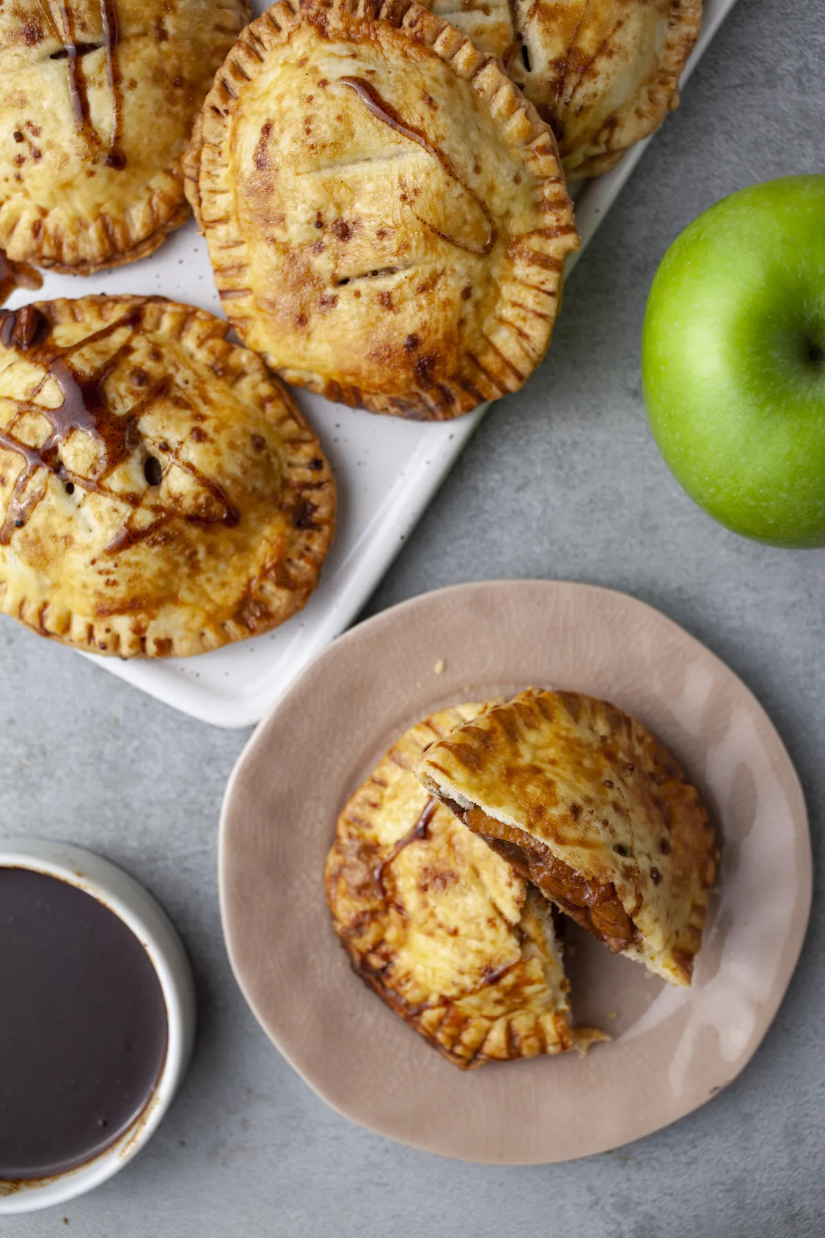 Baked apple hand pies on a serving board, with one pie split open on a pink plate next to caramel sauce