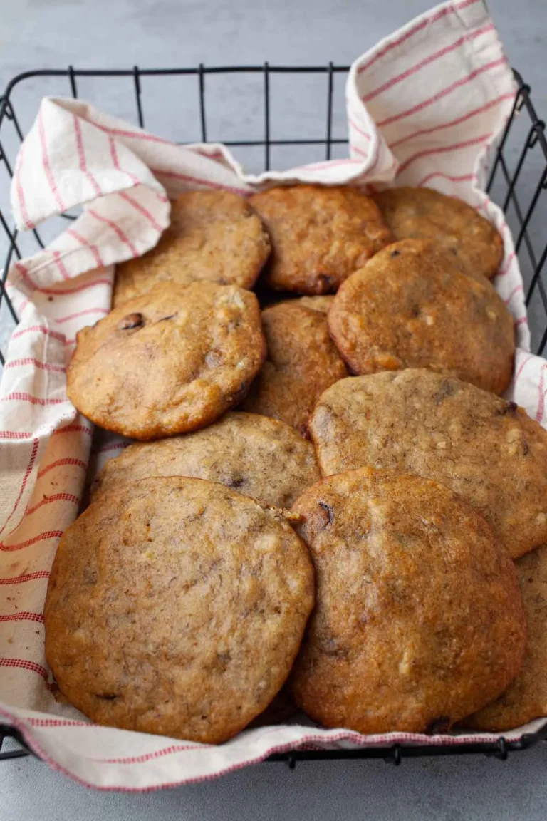 Banana bread chocolate chip cookies piled in a black wire basket lined with a cream and red striped tea towel, golden brown at the edges, on a gray surface.
