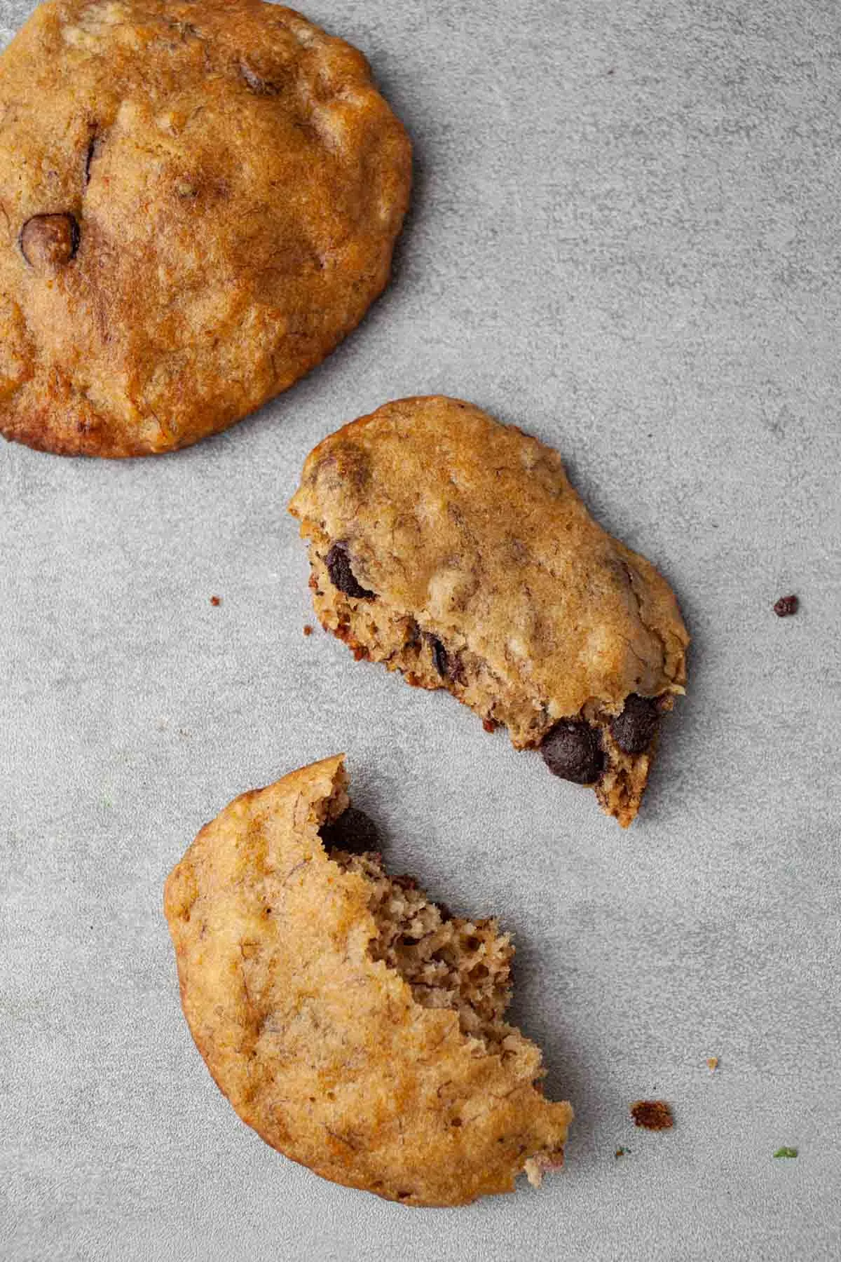Overhead close-up of soft banana bread cookies broken open to show the cakey crumb and chocolate chips, with crumbs scattered on a light gray surface.