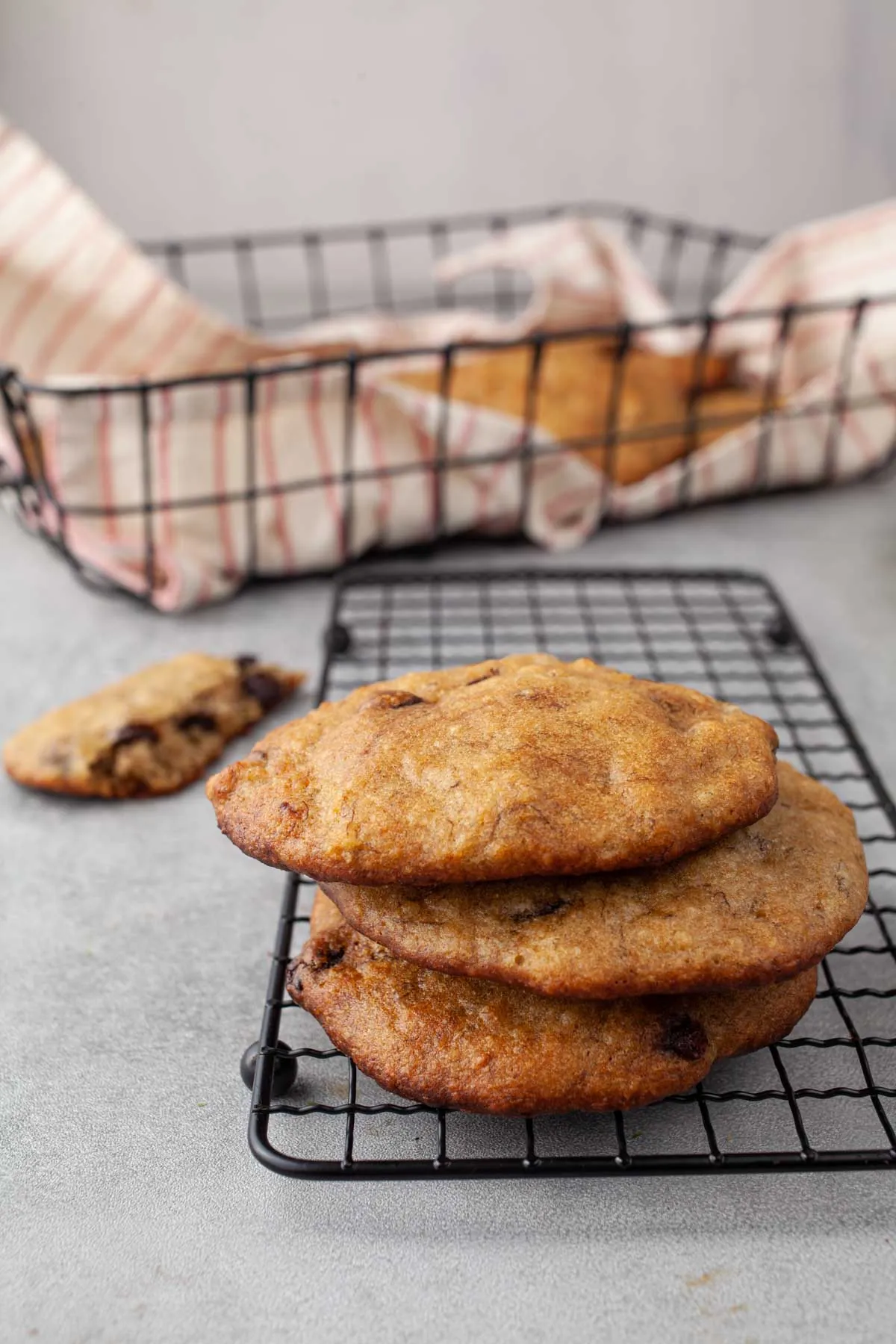 A stack of three banana bread chocolate chip cookies on a black cooling rack, with a broken cookie beside it and a wire basket of cookies blurred in the background.