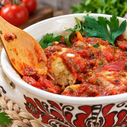 A wooden spoon lifting a beet and lentil ball out of the tomato sauce, showing its soft golden inside, in a painted ceramic bowl with parsley and tomatoes behind.