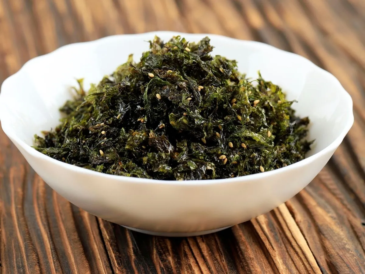 Crumbled toasted nori mixed with sesame seeds in a white scalloped bowl on a dark wooden table, a simple homemade seaweed sprinkle.