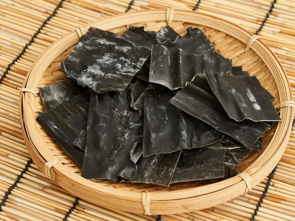 Pieces of dried kombu with a white powdery coating, piled in a round bamboo basket on a bamboo mat.