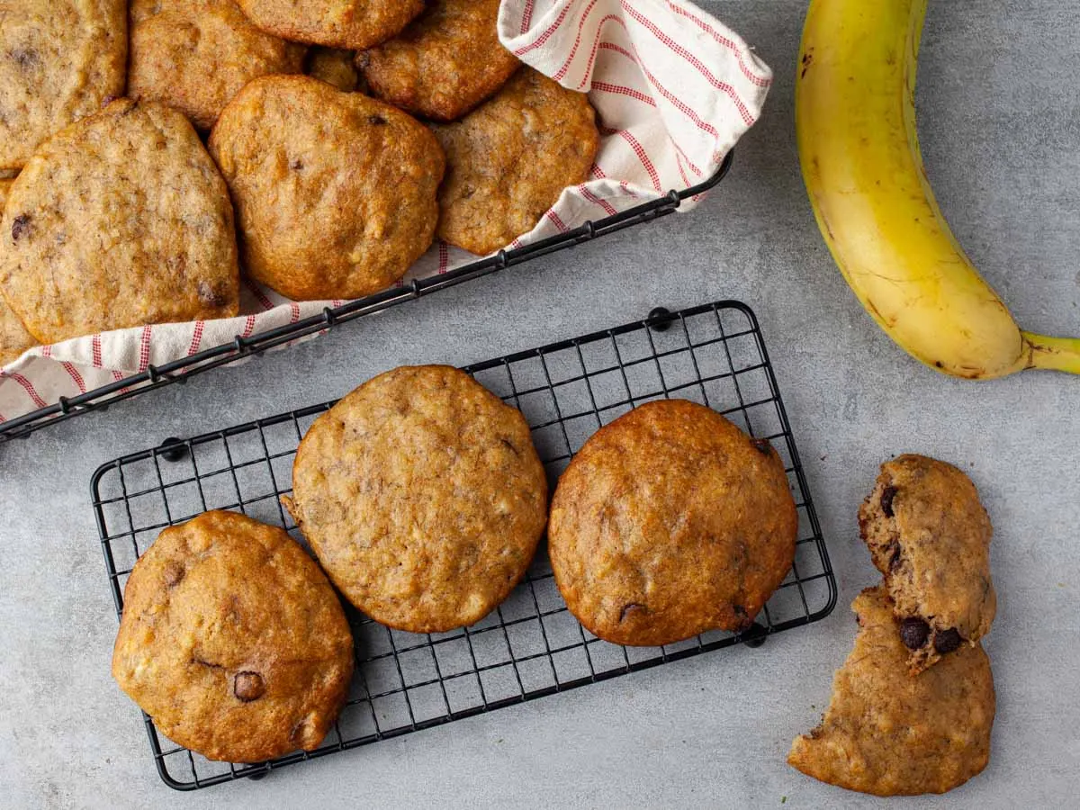 Banana bread chocolate chip cookies cooling on a black wire rack, seen from above, next to a basket lined with a striped tea towel, a ripe banana and a cookie broken in half.