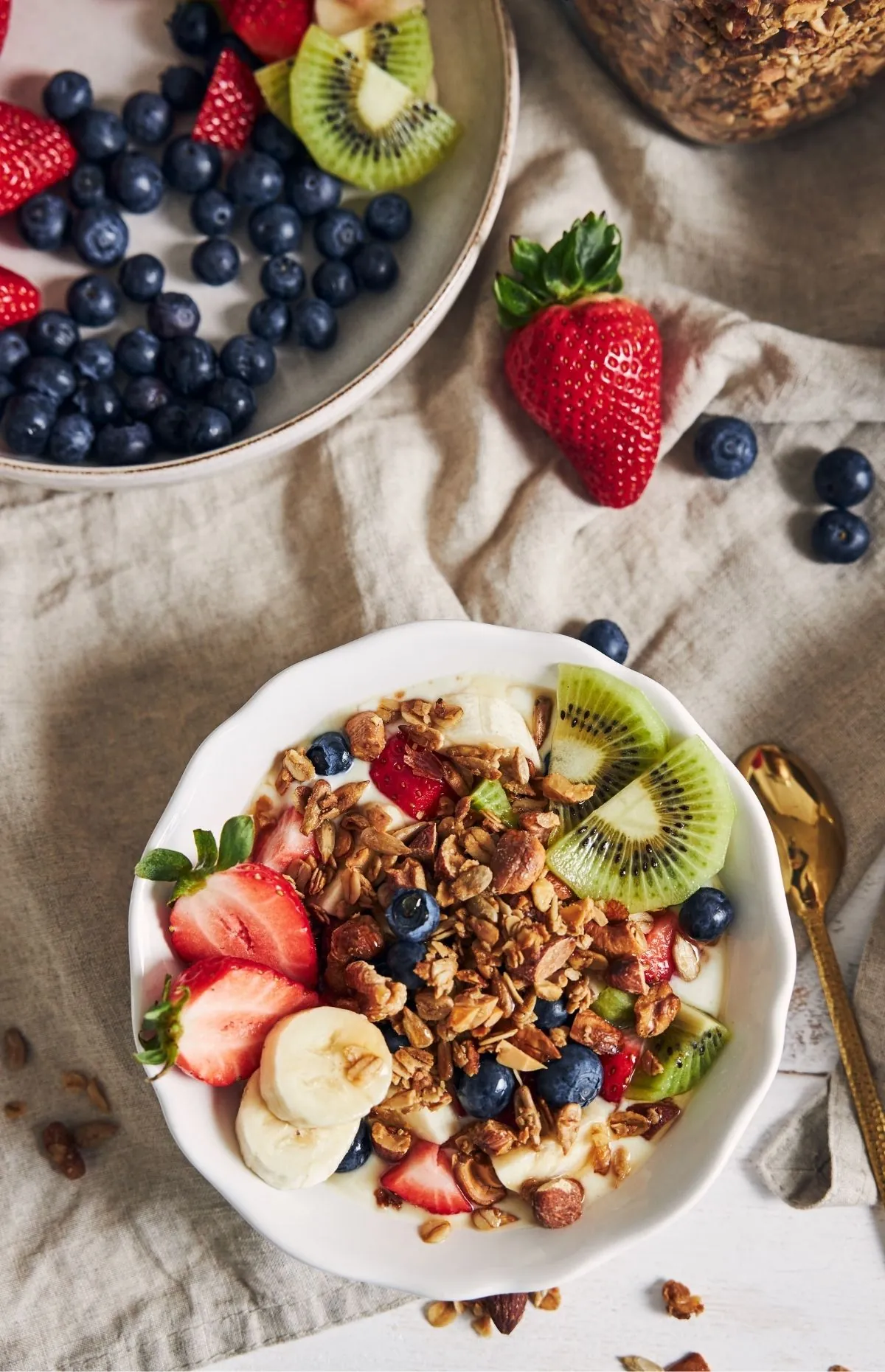 Greek yogurt breakfast bowl topped with crunchy granola, sliced strawberries, kiwi, banana and blueberries, shot overhead on a linen napkin next to a gold spoon and a plate of berries.