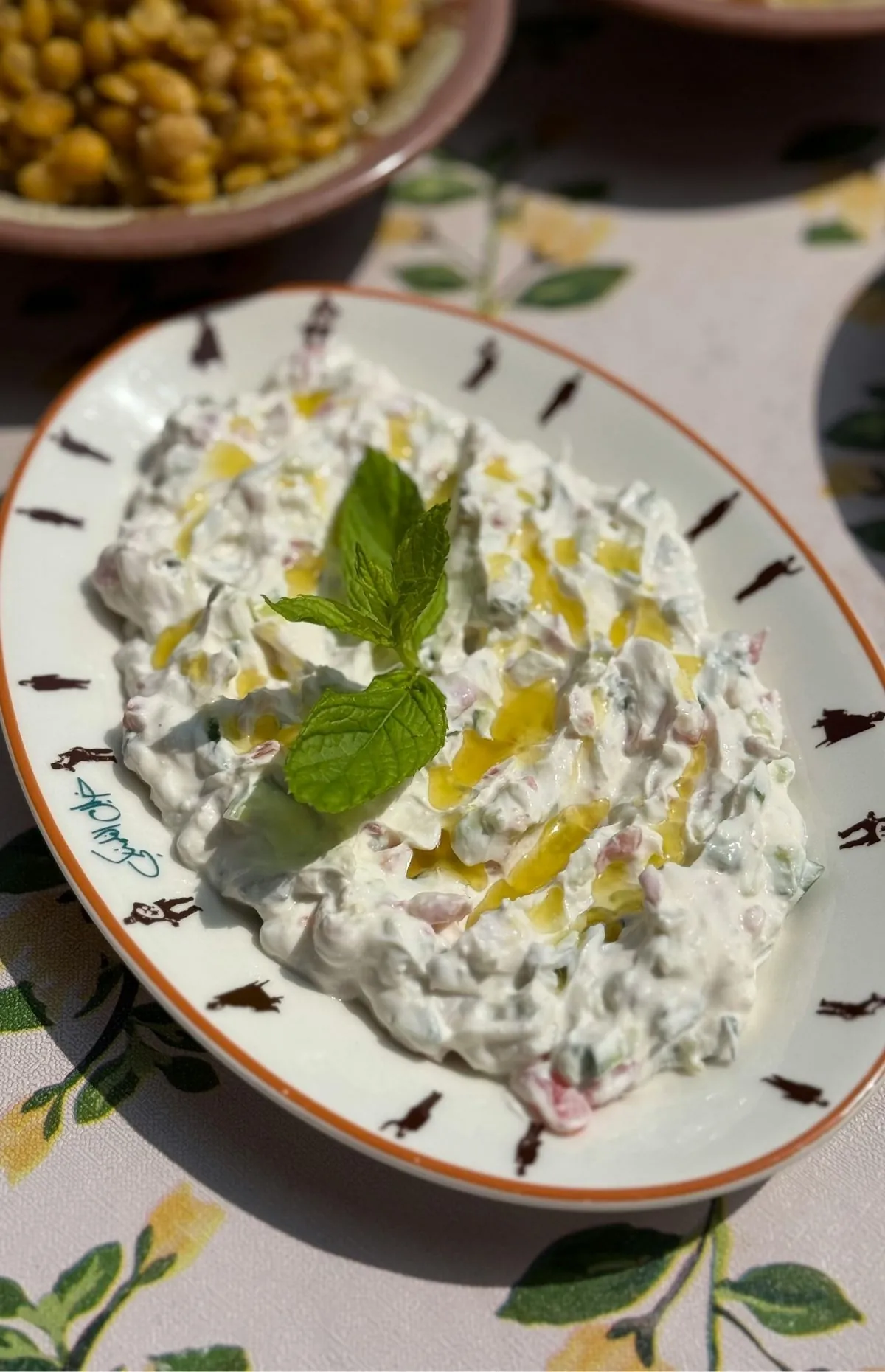 Thick Greek yogurt dip with finely chopped cucumber, drizzled with olive oil and topped with a sprig of fresh mint, served on an oval plate with an orange rim.
