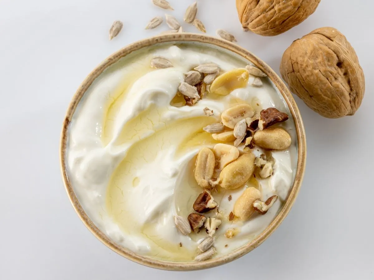 Bowl of plain Greek yogurt swirled with honey and topped with roasted peanuts, chopped pecans and sunflower seeds, shot overhead next to two whole walnuts on a white surface.