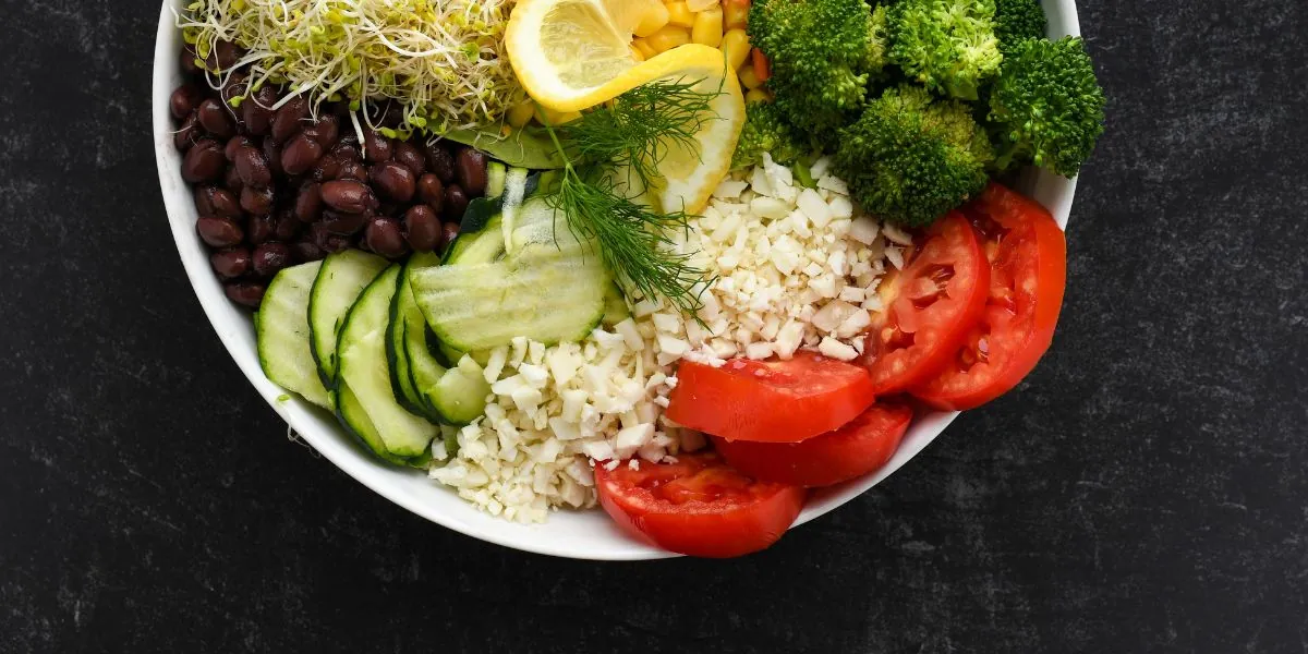 High raw vegan bowl with alfalfa sprouts, broccoli florets, sliced tomatoes, cucumber, riced cauliflower, black beans, dill and lemon on dark slate.