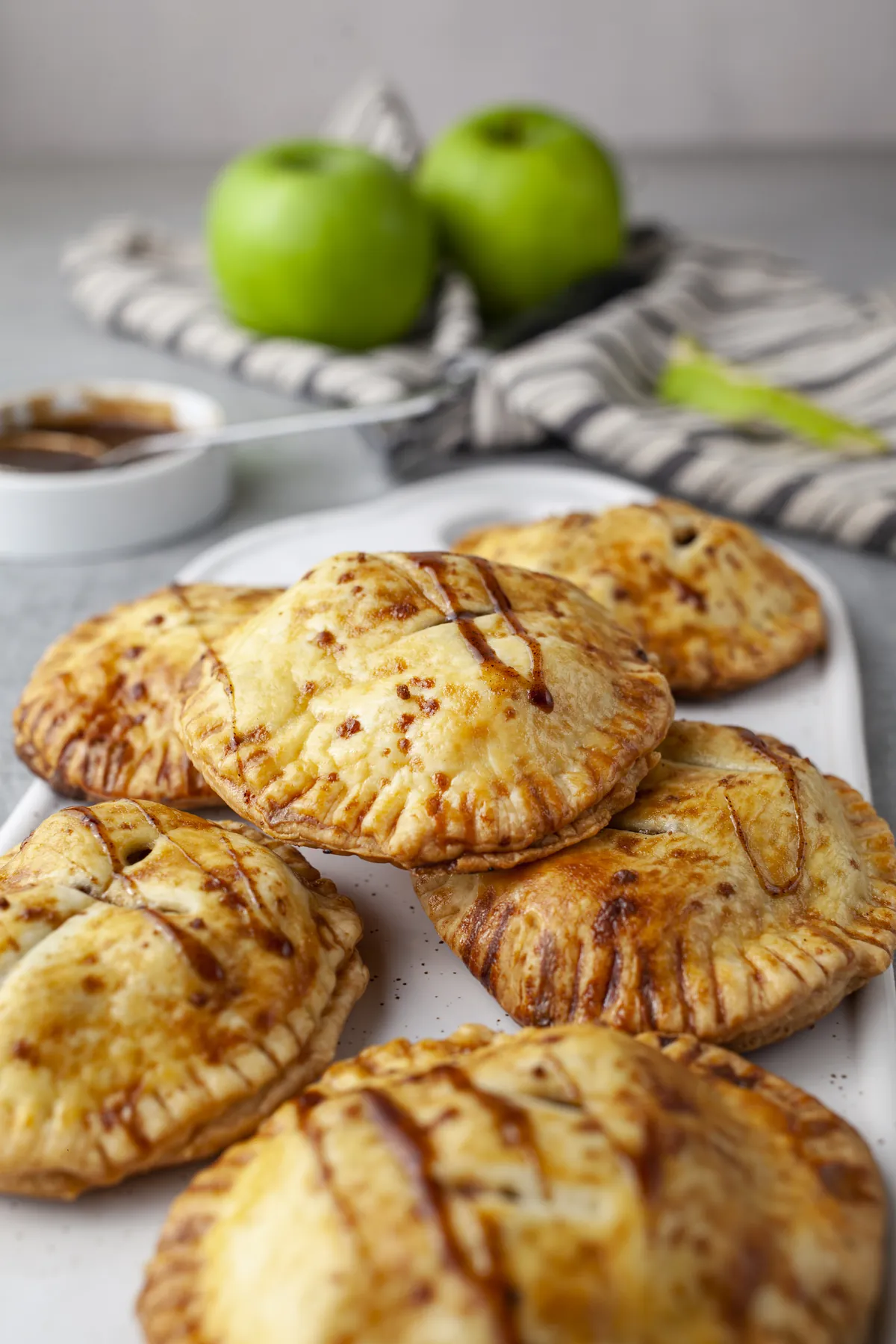 Homemade apple hand pies drizzled with caramel, piled on a white serving board with green apples behind