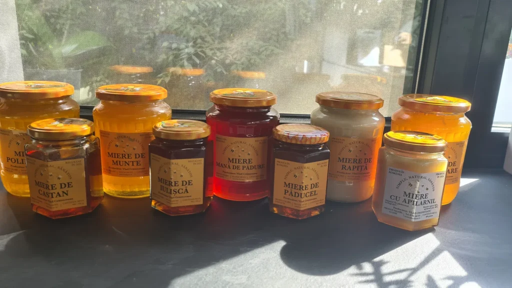 Nine jars of Romanian honey on a sunny windowsill, from pale creamy rapeseed honey to dark knotweed and ruby honeydew honey