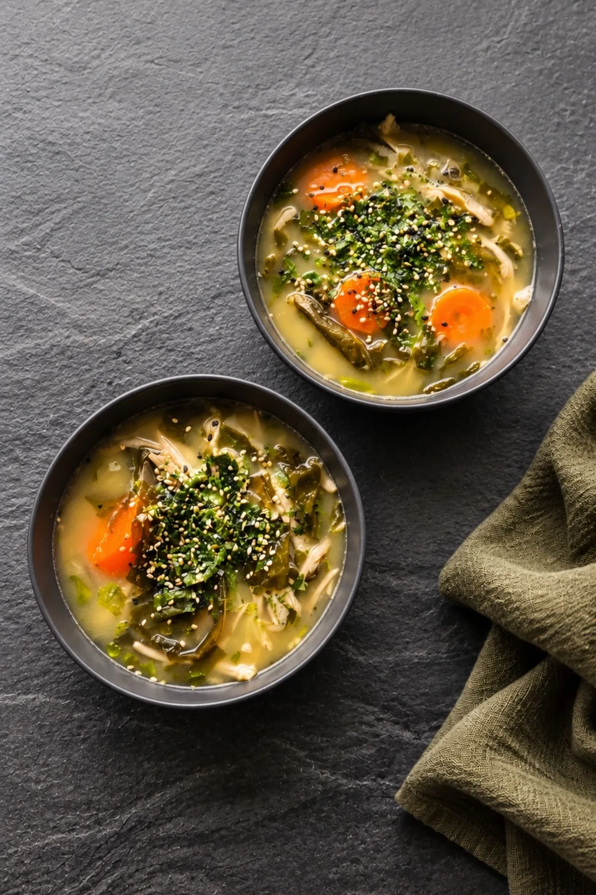 Overhead view of two bowls of miso noodle soup with carrot rounds, wakame and soba, each topped with a mound of sesame and nori furikake, on a dark slate surface with an olive linen napkin.