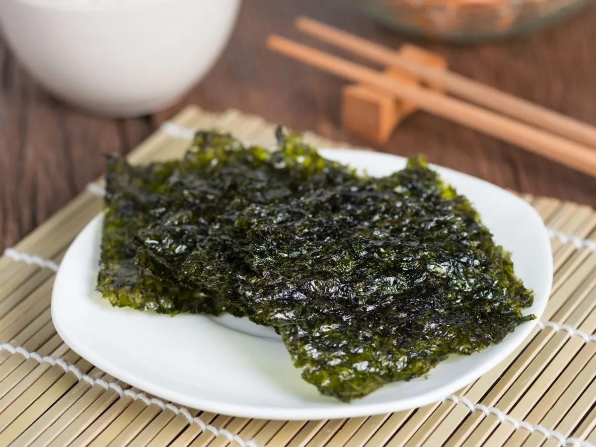Toasted nori sheets on a white plate set on a bamboo sushi mat, with chopsticks and a bowl of rice in the background.