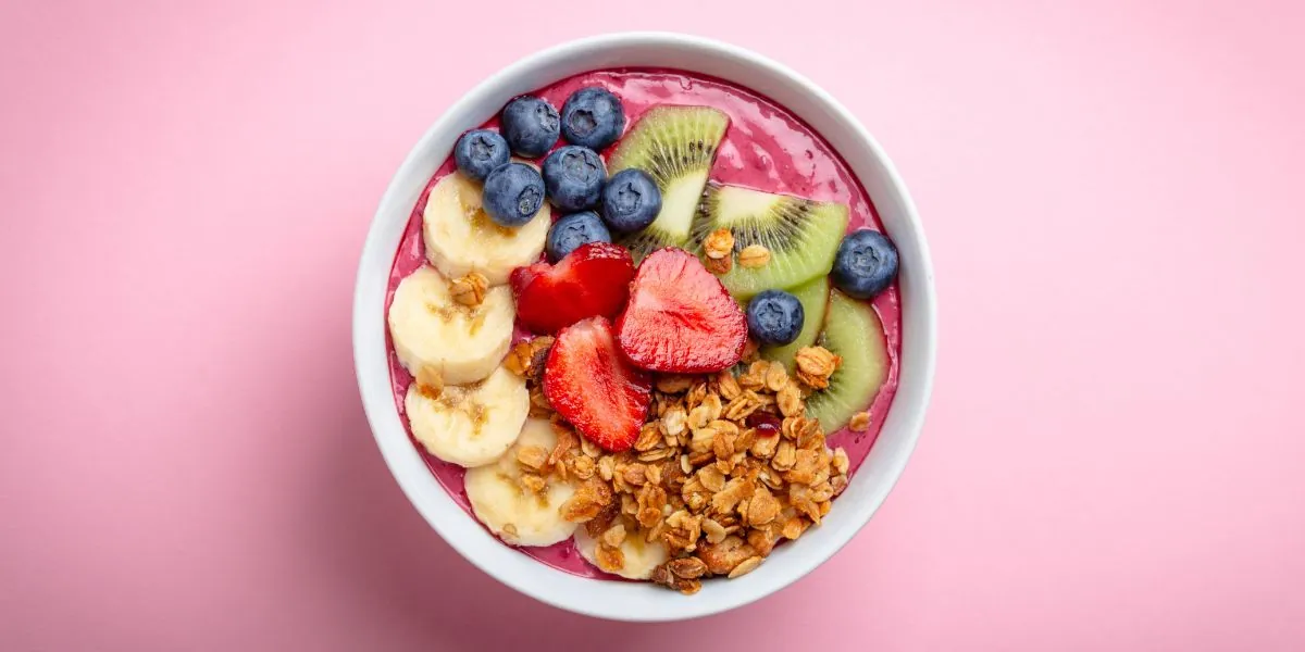 Pink smoothie bowl topped with sliced banana, strawberries, kiwi, blueberries and granola, in a white bowl on a pink background, seen from above.