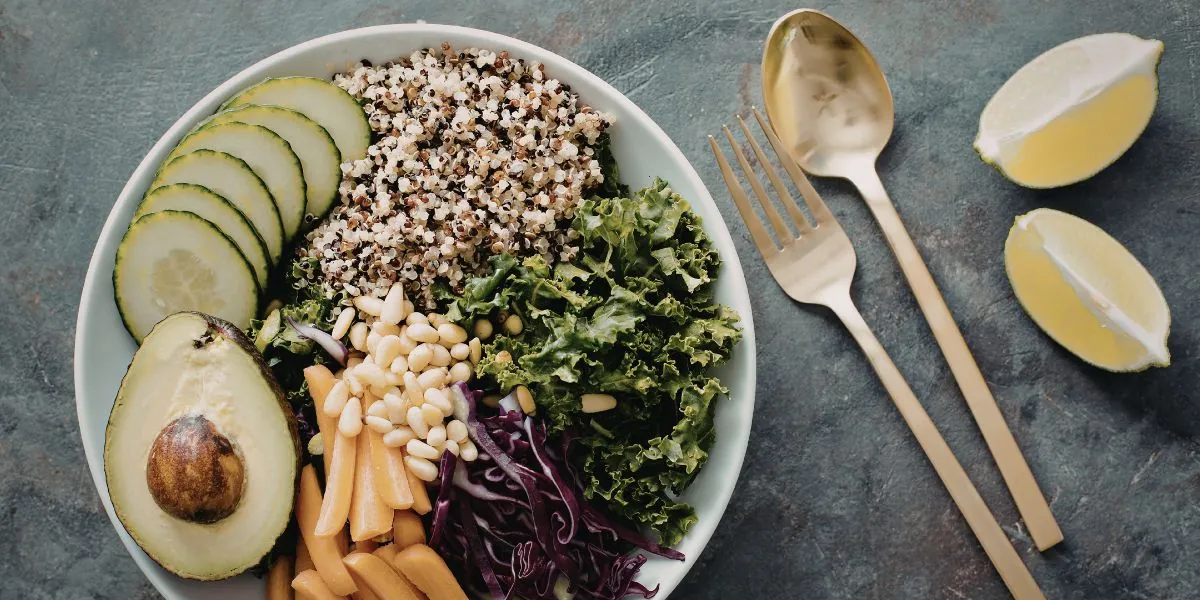 Overhead view of a plant bowl with cucumber slices, tricolor quinoa, kale, half an avocado, pine nuts, carrot sticks and red cabbage, beside gold cutlery and lemon.