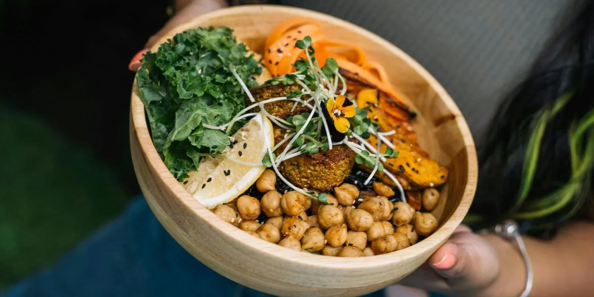 Hands holding a wooden bowl of kale, chickpeas, falafel, roasted sweet potato, carrot ribbons, microgreens and a lemon slice.