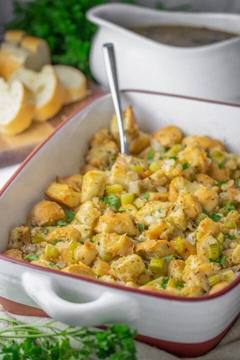 Vegan celery stuffing in a white baking dish with golden bread cubes, celery and parsley, a gravy boat and sliced bread behind.