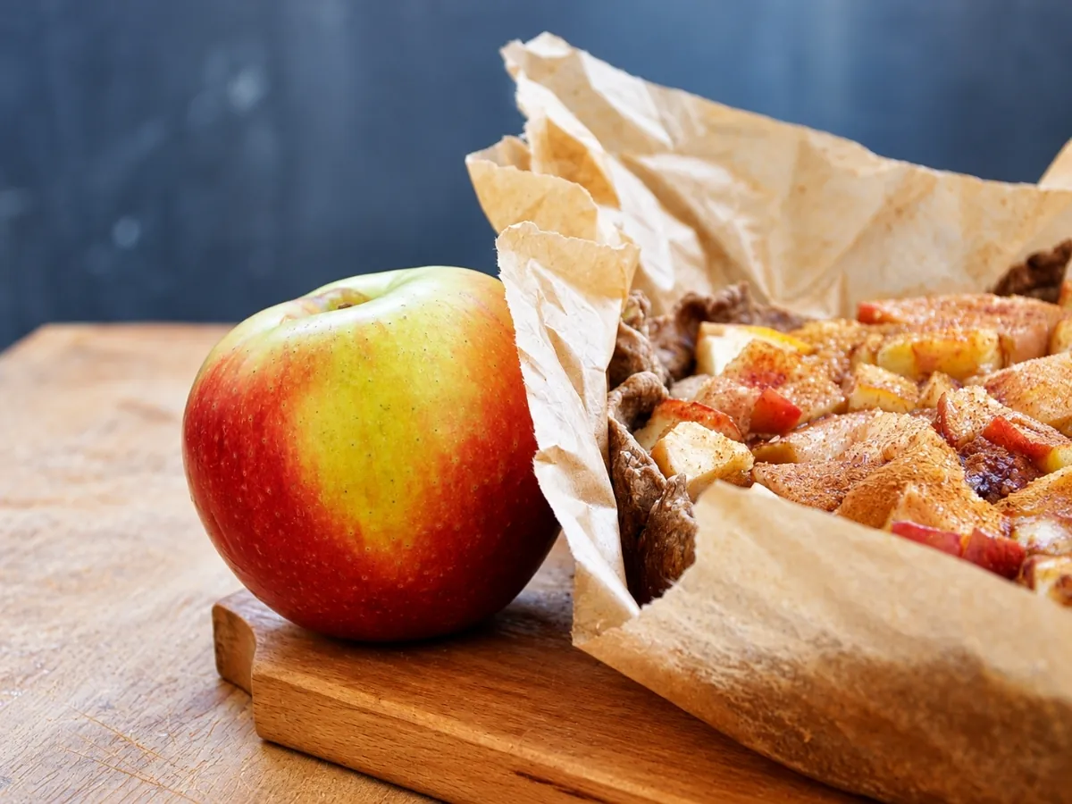Rustic apple tart wrapped in parchment next to a fresh red and yellow apple