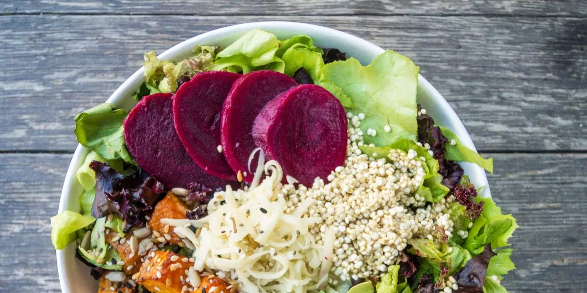 Salad bowl with mixed lettuce, sliced beets, shredded vegetables, puffed grains, squash cubes and seeds, on a weathered blue wooden table.