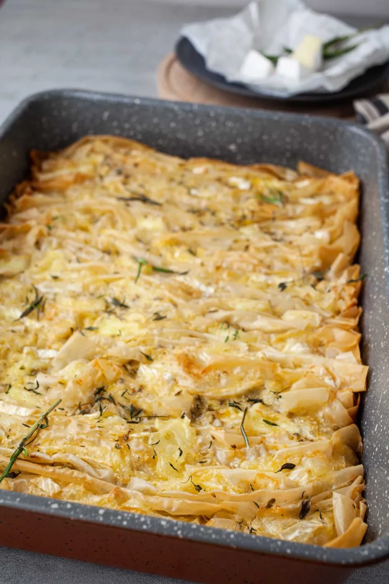 Ruffled phyllo crinkle pie baked until golden, with pockets of melted Camembert and thyme leaves, in a gray baking pan, with Camembert cubes and rosemary on a plate behind it.