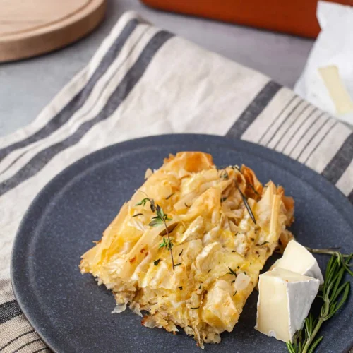 A square slice of Camembert crinkle pie on a dark blue plate, showing crisp phyllo ruffles set in the egg custard, with two Camembert wedges and a rosemary sprig, the pan in the background.