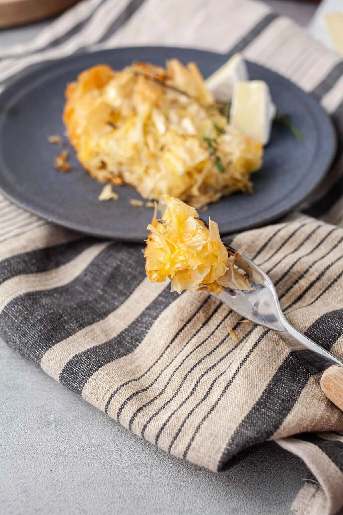 O bucată de plăcintă sărată cu foi încrețite pe furculiță, cu foi fragede și camembert topit, deasupra unui prosop de in în dungi, cu restul feliei pe farfuria albastră.