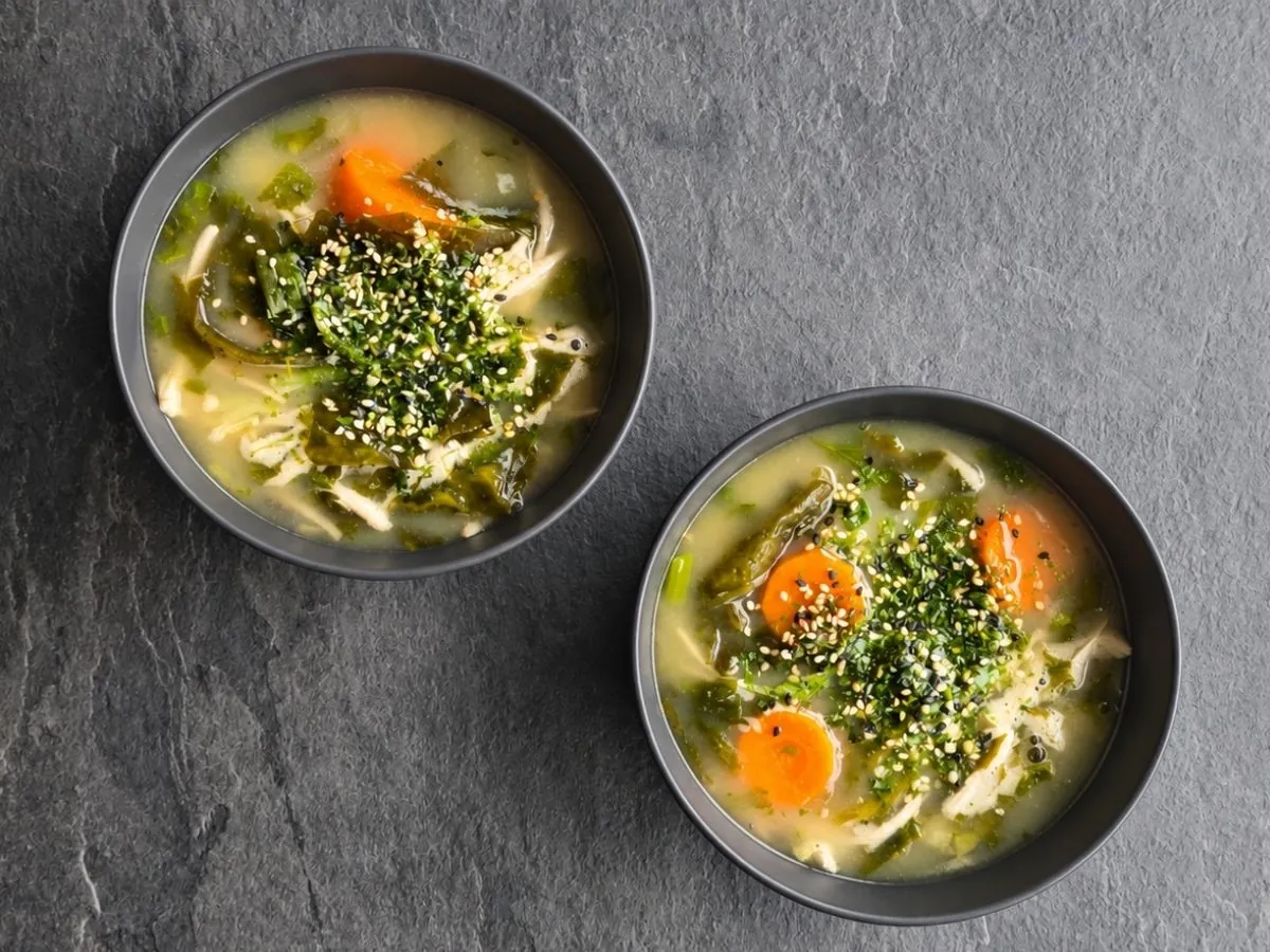 Two bowls of soba soup with wakame, carrot rounds and noodles in a clear golden miso broth, topped with toasted white and black sesame and nori flakes, shot from above on gray slate.