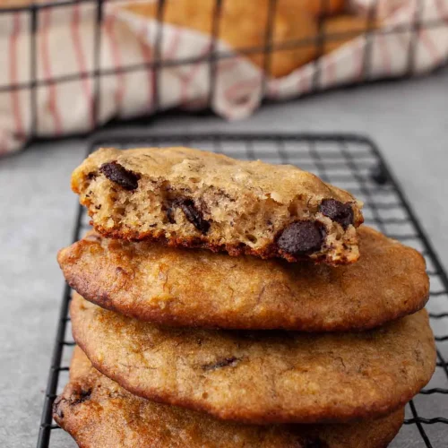 Close-up of stacked banana bread cookies on a wire rack, the top one broken in half to show a soft, cake-like center studded with dark chocolate chips.
