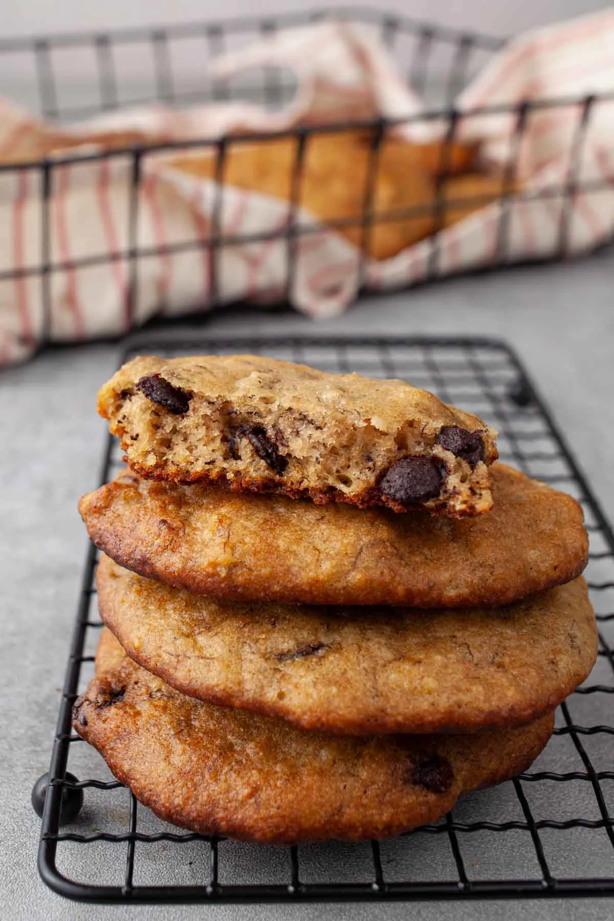 Close-up of stacked banana bread cookies on a wire rack, the top one broken in half to show a soft, cake-like center studded with dark chocolate chips.