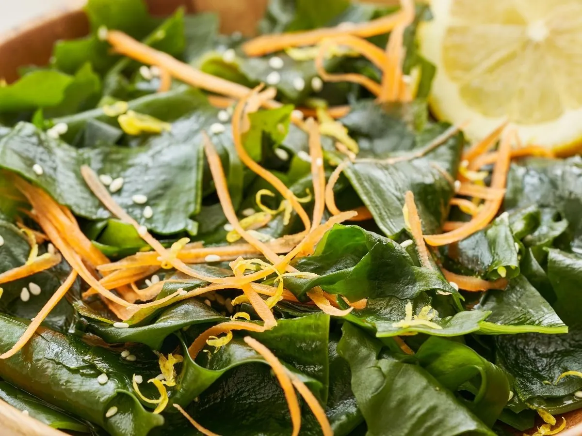 Wakame seaweed salad with thin carrot strips, sesame seeds and lemon zest, close-up, with a lemon slice at the edge of the wooden bowl.