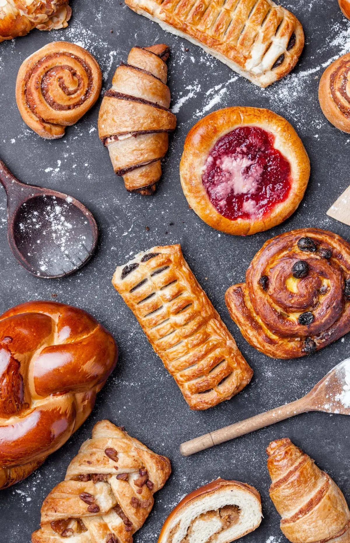 Produse de patiserie din făină albă, cu melci cu scorțișoară, cornulețe, un pateu cu gem, un melc cu stafide și o pâine împletită, pe un blat închis la culoare, presărat cu făină.